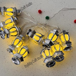May include: A string of decorative silver lanterns with warm yellow lights. The lanterns are arranged on a light-colored surface, with small red and green pom-poms scattered around. The text "ItsyBitsy Craft studio" is visible.