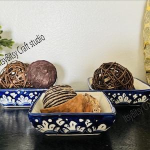 May include: Three blue and white ceramic square bowls with decorative floral patterns. Each bowl is filled with decorative balls made of woven natural fibres.