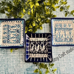 May include: Three square ceramic dishes with intricate blue and white patterns. The dishes have a dark blue border and are arranged on a light blue surface with green foliage. The text "Bitsy Craft studio" is visible.