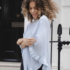 Könnte beinhalten: Ein hellblauer Strickponcho mit Rüschen an den Kanten. Der Poncho wird von einer Person mit lockigem Haar getragen.