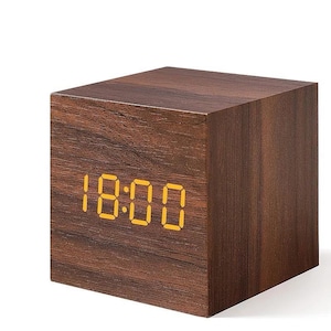 May include: A wooden cube digital clock displaying the time 18:00 in yellow digits. The clock has a dark brown wood grain finish. The cube shape is modern and minimalist.