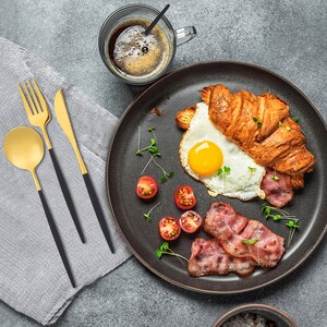 Könnte beinhalten: Ein schwarzer Teller mit einem Croissant, einem Spiegelei, Speck und Kirschtomaten. Der Teller steht auf einer grauen Oberfläche mit einer Tasse Kaffee und Besteck.