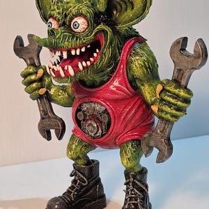May include: A green monster figurine with large ears, red eyes, and a wide mouth holding two wrenches. The monster wears a red tank top and black boots. The figurine has a detailed, textured appearance.