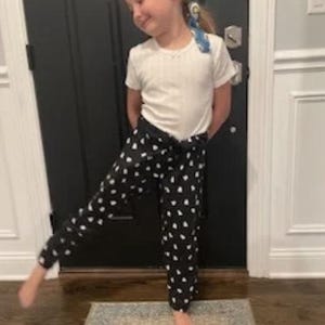 May include: Black pajama trousers with white heart patterns. The trousers are worn with a white t-shirt. The outfit is casual and comfortable, suitable for lounging or sleepwear. The trousers have a drawstring waist.