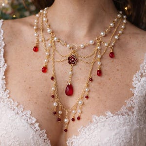 May include: A layered gold-tone necklace with faux pearls and red gemstone accents. The necklace features a central floral pendant and dangling teardrop-shaped gems. The design includes multiple strands and delicate chain details, creating an elegant and ornate look.