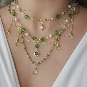 May include: A multi-strand gold necklace featuring green and white beads, pearls, and dangling pendants. The necklace has a starburst charm and a heart-shaped pendant with a green stone. The jewelry is layered and delicate.