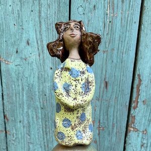 Könnte beinhalten: Ein vertikaler Gartenstecker mit einer handgeformten Keramikfigur mit braunem Haar, die ein gelbes Kleid mit blauen Punkten trägt. Unter der Figur befinden sich eine große, texturierte grüne Kugel, eine zylindrische beige Perle mit Auge und eine rote ovale Perle.