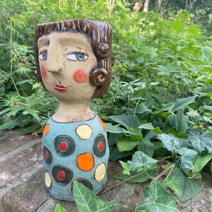 Könnte beinhalten: Ein Keramik-Pflanztopf in Form eines Frauenkopfes mit braunen Haaren, einem blau-weiß gepunkteten Kleid und einem bemalten Gesicht.