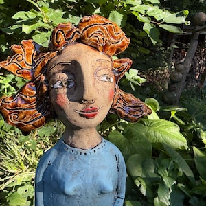 Könnte beinhalten: Eine Keramikskulptur einer Person mit stilisiertem, glänzendem braunem Haar, das dunkle Wirbelmuster aufweist. Die Figur hat ein hellbraunes Gesicht mit bemalten roten Lippen und rosigen Wangen und trägt ein strukturiertes hellblaues Oberteil.