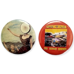 Puede incluir: Dos pines redondos con forma de bot&oacute;n que muestran el arte de la portada del &aacute;lbum de la banda Neutral Milk Hotel. El primer pin muestra a una mujer con un parche de tambor como cabeza, de pie en un barco con un ni&ntilde;o. El segundo pin tiene un fondo rojo con el t&iacute;tulo del &aacute;lbum "Neutral Milk Hotel" y el nombre del &aacute;lbum "In the Aeroplane Over the Sea" en texto blanco.