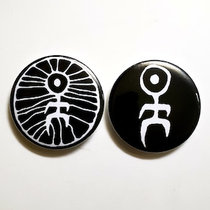 May include: Two black and white round buttons. One button features a white stylized sunburst design with a central figure. The other button has a white abstract figure on a black background. Both buttons are on a white surface.