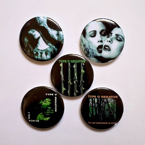 May include: Four black and green round buttons with the band name "Type O Negative" printed on them. The buttons feature different images, including a woman with long black hair, two women with their faces close together, a black background with green thorns, and a black and green forest scene.