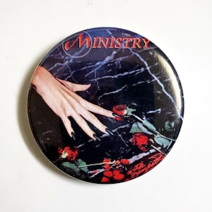 May include: A black and white button featuring a hand reaching towards red roses with the word "MINISTRY" in red lettering.