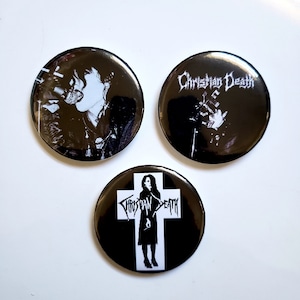 May include: Three black and white button pins with the band name "Christian Death" printed in white. One pin features a black and white image of a person singing into a microphone. Another pin features a black and white image of a person holding a microphone. The third pin features a black and white image of a person standing in front of a cross.