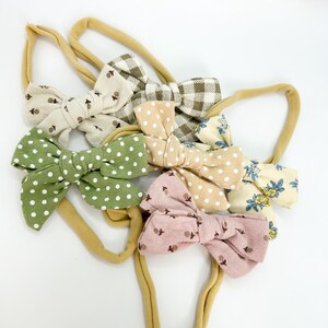 May include: Set of six fabric headbands with bows in various patterns, including gingham, polka dots and floral prints. The headbands are a light brown colour.