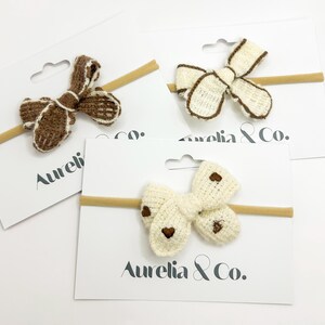 May include: Three knitted baby headbands with bow details. One bow is brown with white edging, another is cream with brown edging, and the third is cream with brown heart details. Each bow is attached to a tan elastic band and displayed on a white card with the text "Aurelia & Co."