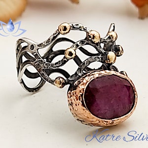 Puede incluir: Un anillo de plata con una piedra preciosa grande, ovalada, de color rojo oscuro engastada en un engaste de oro. El anillo tiene un diseño delicado e intrincado con acentos dorados.