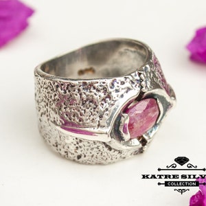 May include: A silver ring with a textured band and a pink gemstone set in a claw setting. The ring is made by Katre Silver Collection.