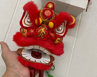 May include: A red and gold Chinese lion dance head with a red tassel. The head is made of fabric and has a white face with black eyes and a red nose. The head is open and the mouth is showing teeth.