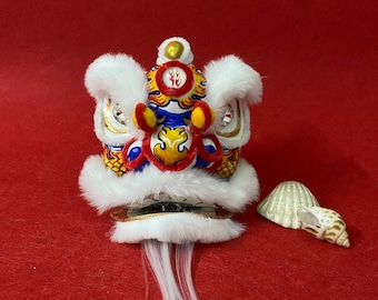 May include: A white and red lion dance head with gold accents and a white fur mane. The head is decorated with blue, yellow, and red paint. The head is on a red background.