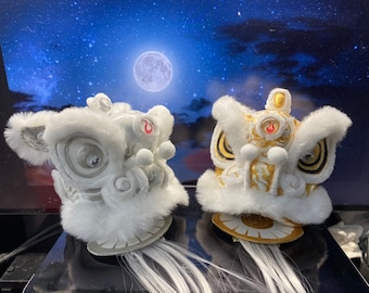 May include: Two white lion dance heads with gold accents. One head has a silver design and the other has a gold design. Both heads have white fur and are on a black surface.