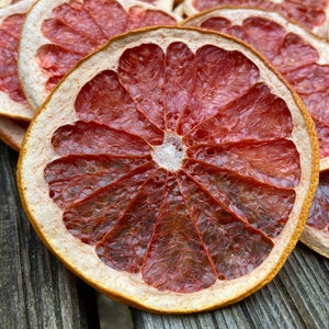 Può includere: Fette di pompelmo essiccate, un prodotto alimentare naturale, disposte su una superficie di legno. Le fette sono di un colore arancione vibrante con una tonalità rossastra, e la texture è traslucida e leggermente rugosa.