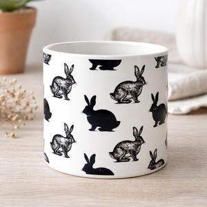 Può includere: Vaso in ceramica bianca decorato con illustrazioni di conigli neri. Il design presenta sia disegni dettagliati che sagome nere di conigli. Il vaso cilindrico è adatto per piante da interno, circa 15 cm di diametro.