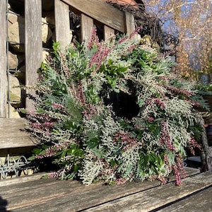 Peut inclure: Une couronne décorative composée de diverses plantes vertes, dont des branches de conifères, et ornée de petites fleurs rouge-violet. La couronne est posée sur un banc en bois vieilli, avec un arrière-plan extérieur naturel.