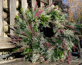 Könnte beinhalten: Ein dekorativer Kranz aus verschiedenen grünen Pflanzen, darunter immergrüne Zweige, und mit kleinen, rot-violetten Blüten verziert. Der Kranz liegt auf einer rustikalen Holzbank, vor einer natürlichen Außenkulisse.