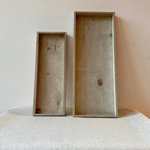 Puede incluir: Dos bandejas de madera, una más grande que la otra, ambas con un acabado gris claro. Las bandejas están hechas de madera con un patrón de veta natural. Están sobre un mantel de lino beige claro.