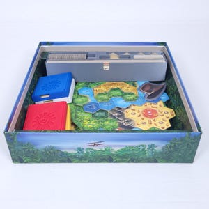 Op de afbeelding: Een spelbord met een blauw, groen en geel ontwerp. Het bord is omgeven door een groene en bruine rand. Er zijn verschillende kleine, gekleurde dozen en een klein vliegtuig op het bord.