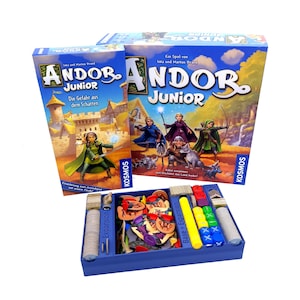 Könnte beinhalten: Ein Brettspiel-Box-Set mit dem Titel "Andor Junior" in Blau und Gelb. Die Box zeigt Illustrationen von Fantasy-Charakteren und einer Burg. Die Spielkomponenten sind in einer blauen Schale sichtbar, einschließlich Spielsteinen und Würfeln.
