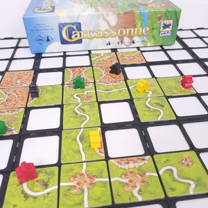 Könnte beinhalten: Das Brettspiel "Carcassonne" mit der Spielschachtel im Hintergrund. Das Spielbrett besteht aus grünen Kacheln mit Straßen und Gebäuden. Holzfiguren in Rot, Gelb, Grün und Schwarz sind auf dem Spielbrett platziert. Das Spiel ist aufgebaut und spielbereit.