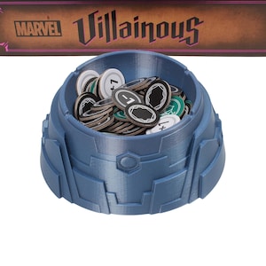 Puede incluir: Un porta fichas de plástico azul lleno de fichas redondas con números negros y diseños verdes y negros. El porta fichas está etiquetado como "Villainous" en texto negro sobre un fondo marrón. El texto "MARVEL" también es visible en el fondo.