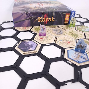 May include: The board game "Karak" is displayed, with a box showing a fantasy scene. The game board is made of hexagonal tiles, with character tokens and game pieces. The game is for 2-5 players aged 10 and over.
