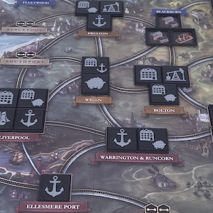 以下が含まれることがあります： 都市の名前とトークンが書かれたゲームボード。トークンには、船、錨、建物、鉱山カート、石油掘削リグが含まれています。ボードは、暗いグレーの素材で、明るいグレーのグリッドパターンが施されています。