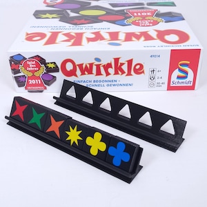 Könnte beinhalten: Ein schwarzer Plastik-Spielsteinhalter mit einer Reihe bunter Spielsteine. Die Steine sind rot, grün, gelb und blau mit einem Stern, einem Kreis und einem Pluszeichen. Der Halter ist mit "Qwirkle" beschriftet und hat ein Spielbrett im Hintergrund.