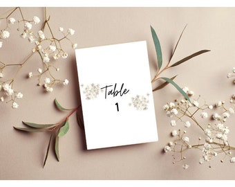 Könnte beinhalten: Weiße Karte mit schwarzem Text "Tisch 1" umgeben von weißen Gipskrautblüten und grünen Eukalyptusblättern auf einem hellbraunen Hintergrund.