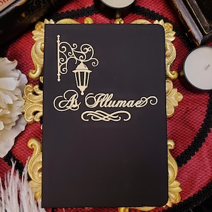 Peut inclure: Un carnet noir avec un motif de lampadaire de rue doré et les mots "As Illumae" en écriture décorative.