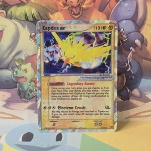 May include: A Zapdos ex trading card with 110 HP, featuring a yellow electric bird illustration, the Legendary Ascent Poke-Power, and the Electron Crush attack. The card has a holographic border and text detailing game mechanics and damage.
