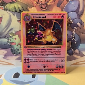 May include: A holographic Charizard trading card featuring the character in a dynamic pose with wings spread. The card displays 120 HP, the Flame Pokémon classification, Energy Burn power, and the Fire Spin attack with 100 damage points.