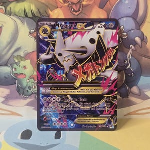 May include: A Mega M Aggron EX trading card with 240 HP, featuring a metallic silver and black creature illustration, pink energy effects, and the attack Megaton Slam. The card includes Japanese text, the Mega Evolution rule, and game statistics.