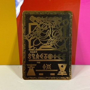 May include: A gold-colored metal card with a black background. The card features a stylized image of a creature with a sword and shield. The text "NAME SKELETON" is printed on the card.