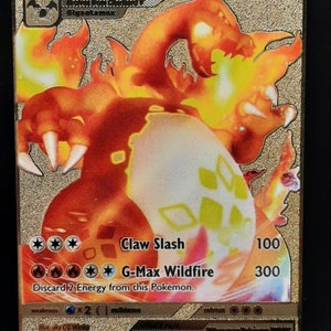 May include: A gold foil Pokemon trading card featuring Charizard VMAX with a fiery orange and yellow design. The card has a large image of Charizard in its G-Max form, with the text "Charizard VMAX" and "330" printed on the card. The card has two attacks: "Claw Slash" for 100 damage and "G-Max Wildfire" for 300 damage. The card also has the text "Discard 2 Energy from this Pokemon."