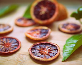 May include: Close-up of dried blood orange slices on a wooden surface with green leaves. The orange slices are a deep red color and have a slightly wrinkled texture.