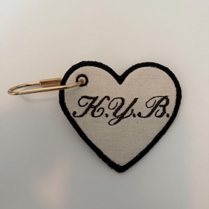 May include: Heart-shaped tag with a black border and the embroidered letters "H.Y.B." in brown. The tag is attached to a gold-colored metal clasp.
