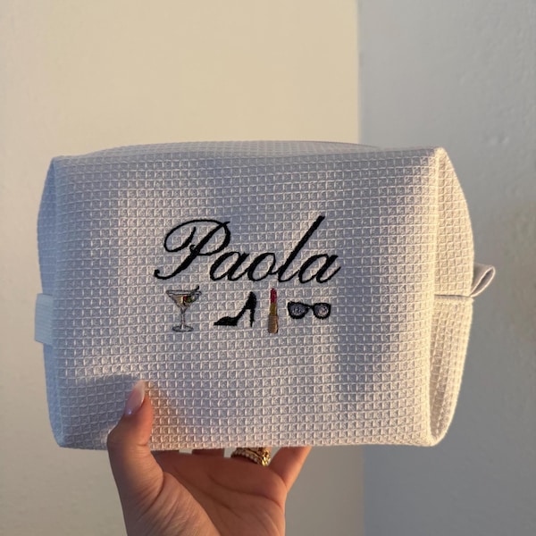 May include: A white waffle-textured cosmetic bag featuring the embroidered name Paola in black script. Below the name are small embroidered icons of a martini glass, a black high-heeled shoe, a red lipstick, and black sunglasses.