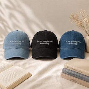 Könnte beinhalten: Drei Baseballkappen in Blau- und Schwarztönen, jede mit dem gestickten Text "I'm not ignoring you, I'm reading." Die Kappen werden mit Büchern wie "The Art of Reading" und "A Month in Paris" gezeigt.
