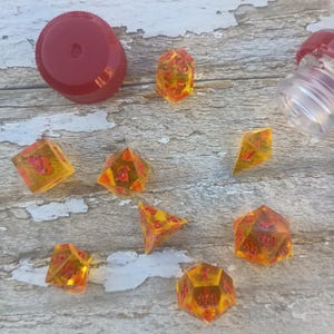 Puede incluir: Un conjunto de ocho dados de &aacute;mbar transl&uacute;cidos con n&uacute;meros rojos. Los dados est&aacute;n esparcidos sobre una superficie de madera clara.