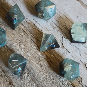May include: Set of seven clear resin dice with blue and white numbers. The dice are in various shapes, including a d4, d6, d8, d10, d12, and d20. The dice are on a wooden surface.